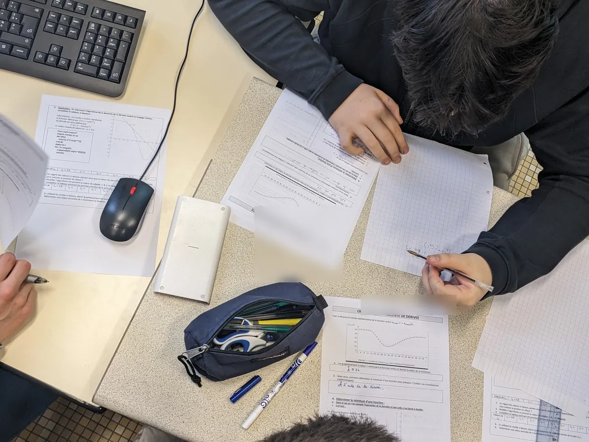 Vue de dessus d’une table d’élèves : fiches d’exercices avec courbes, programme Python et calculs manuscrits.